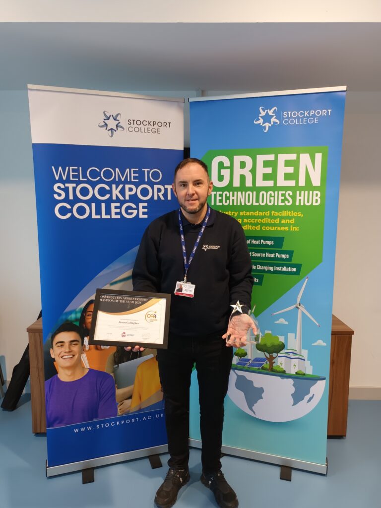 Jason Gallagher holding his trophy and certificate in front of the Stockport College and Green Technologies hub banners.