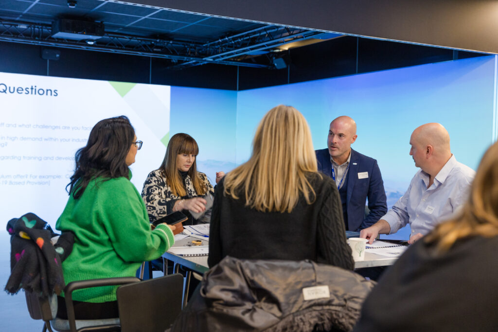 A group of six professionals sit around a table engaged in discussion during a workshop or meeting. Two men in business attire and four women, one wearing a bright green sweater, are talking and taking notes. A presentation slide with the heading “Questions” is projected on the wall behind them, in a modern conference room with soft lighting and a digital screen backdrop.