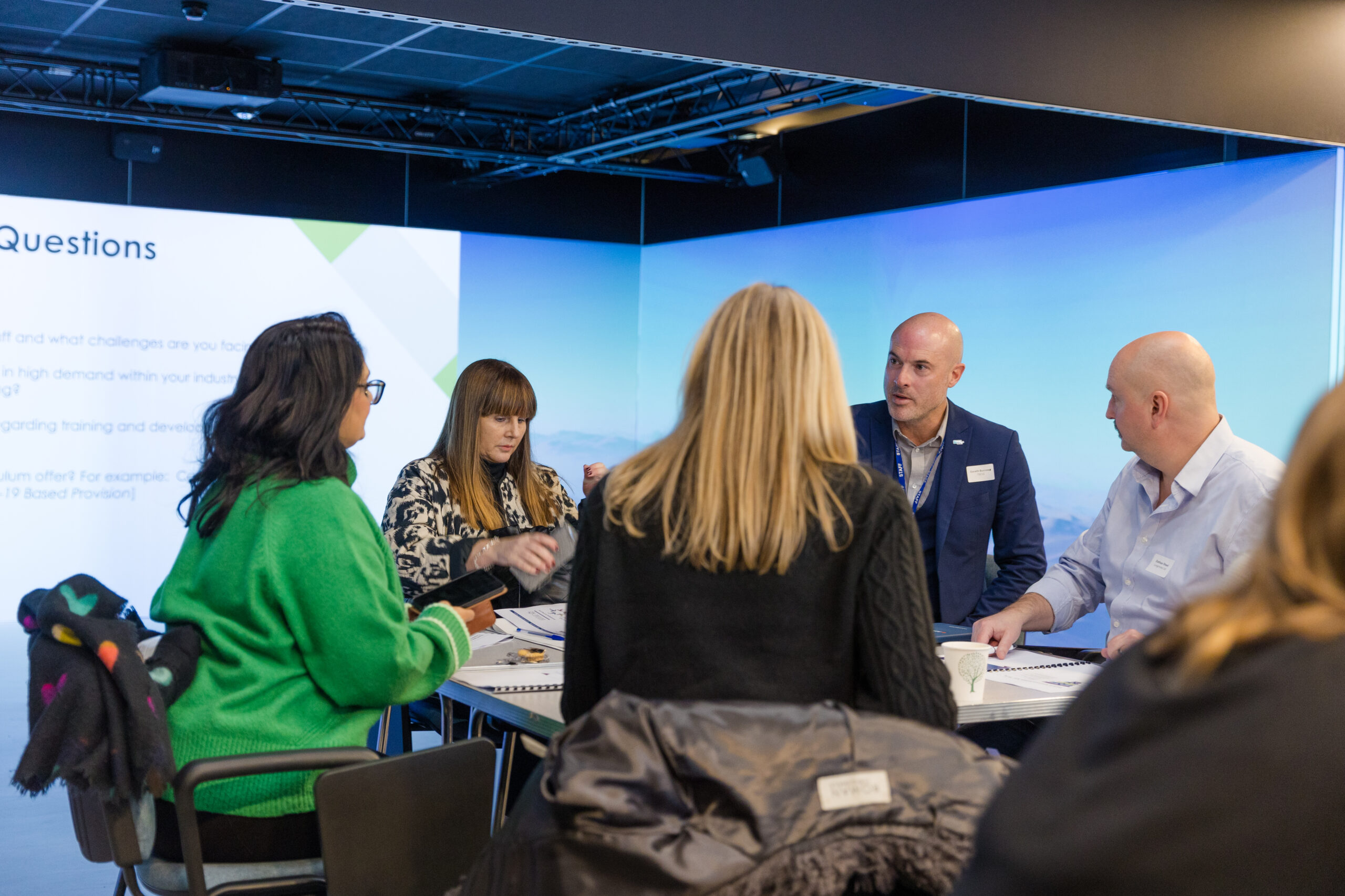 A group of six professionals sit around a table engaged in discussion during a workshop or meeting. Two men in business attire and four women, one wearing a bright green sweater, are talking and taking notes. A presentation slide with the heading “Questions” is projected on the wall behind them, in a modern conference room with soft lighting and a digital screen backdrop.