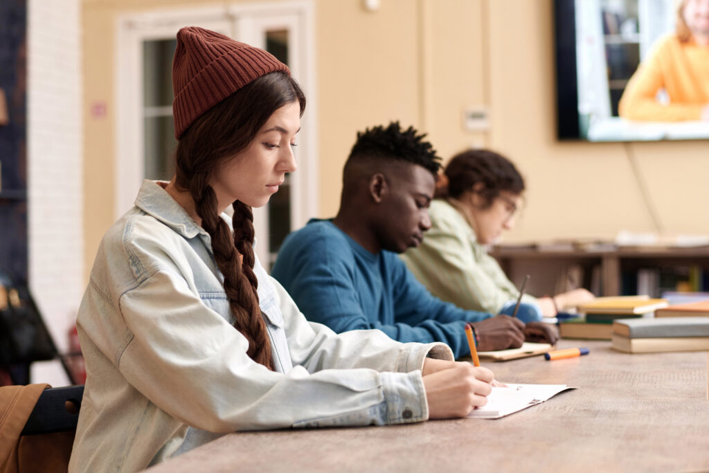 Side view portrait of young woman wearing beanie hat and taking notes during online lecture with teacher on screen in background copy space